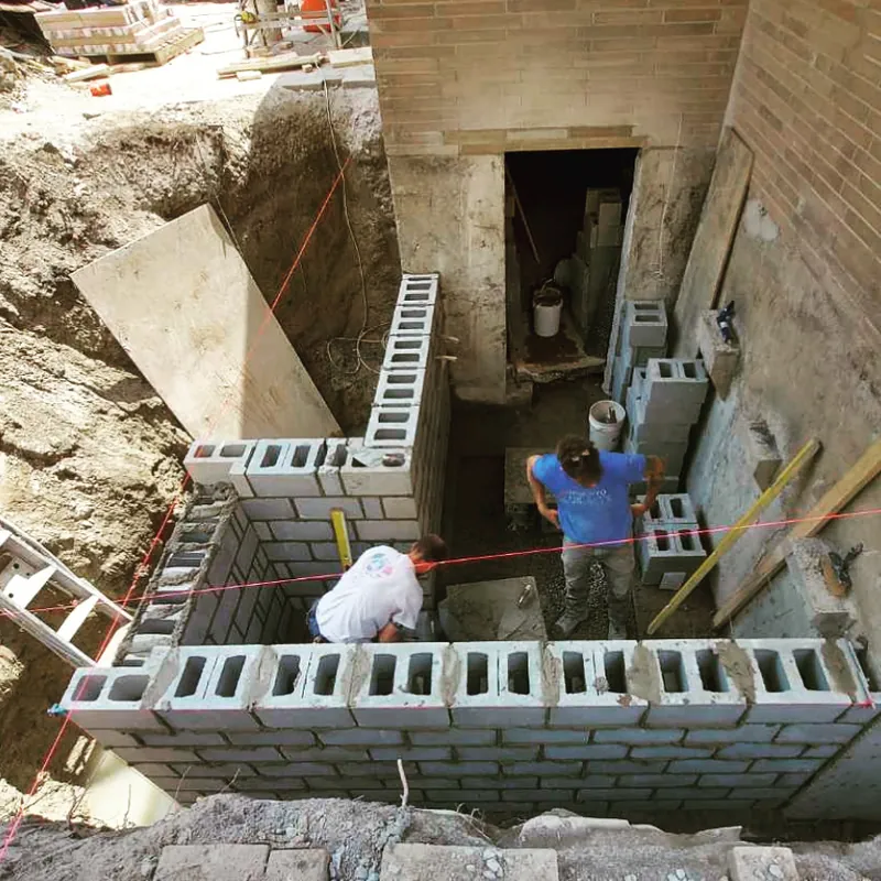 Crew laying a concrete block foundation in an excavation beside a brick building